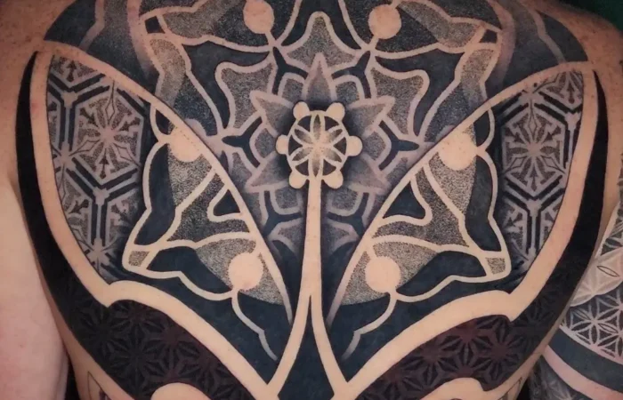 Geometric Tattoos A large-scale geometric back tattoo featuring an intricate mandala design at the center with symmetrical patterns radiating outward. The black ink work showcases detailed dotwork technique creating depth and shading throughout the design. The tattoo covers most of the person's back, displaying precise linework, geometric shapes, and interconnected patterns that demonstrate the technical skill required for this style of body art.
